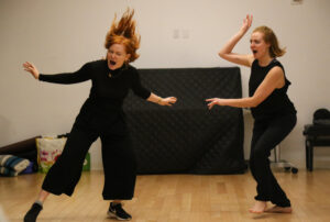 Two women are dancing in a room with black furniture.