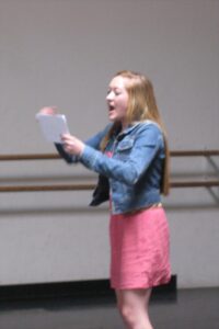 A girl in pink skirt and jean jacket holding paper.
