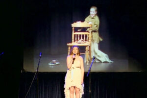 A woman standing on stage with an image of a chair behind her.