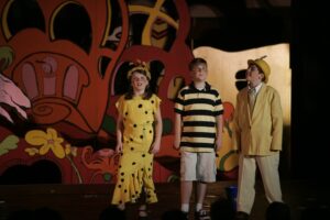 Three children in costumes on stage with a painting behind them.